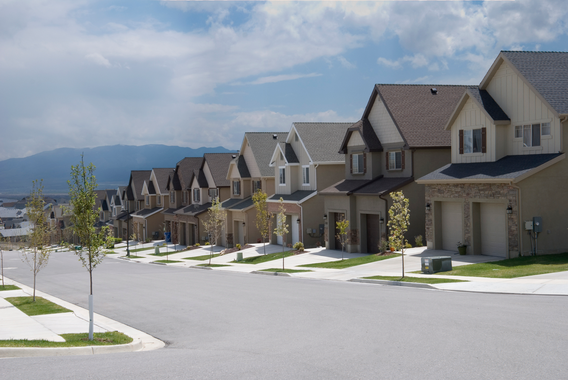 Suburban street with modern houses, sidewalks, and distant mountains under a cloudy sky