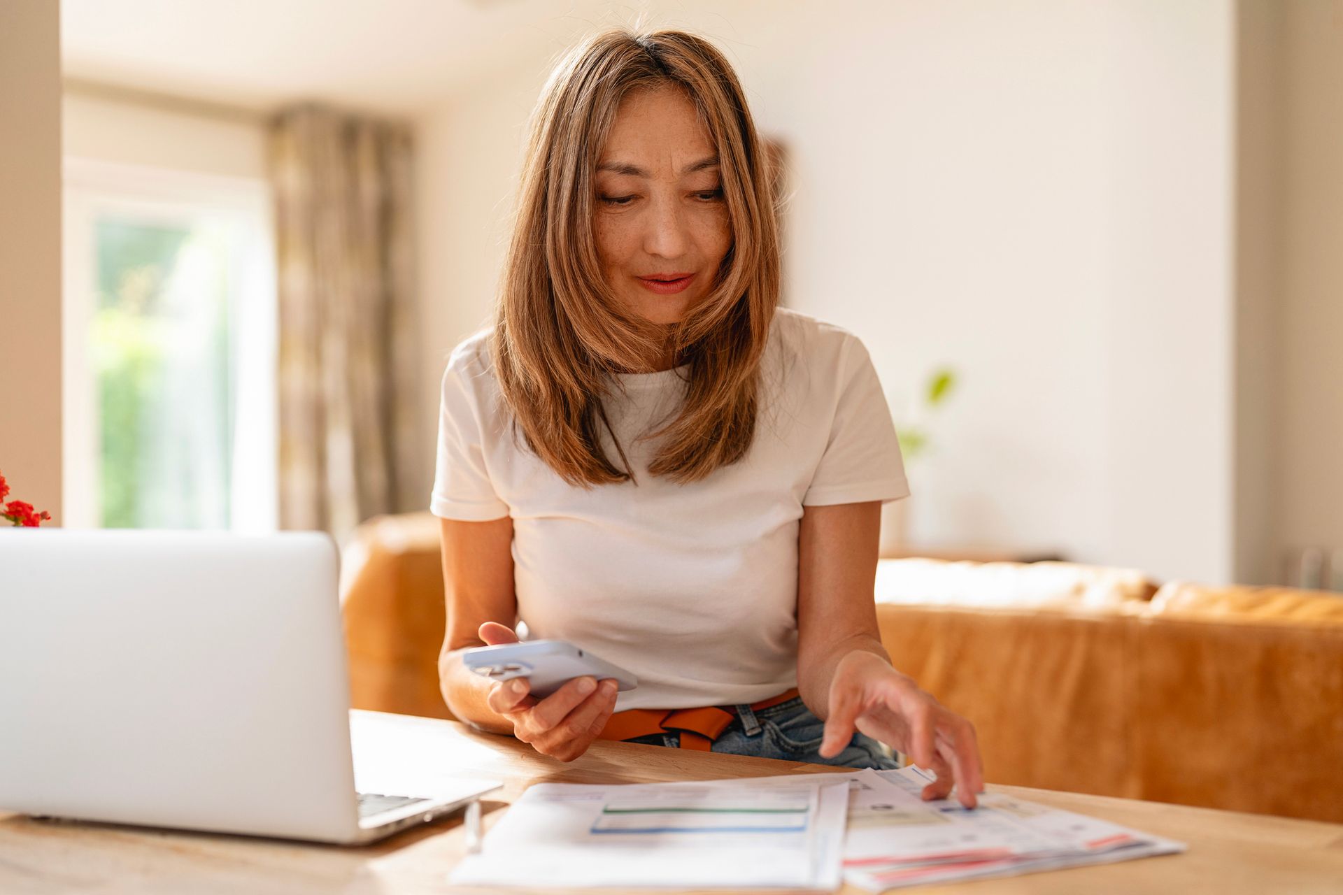 Woman using a smartphone beside a laptop at a desk in a bright home office