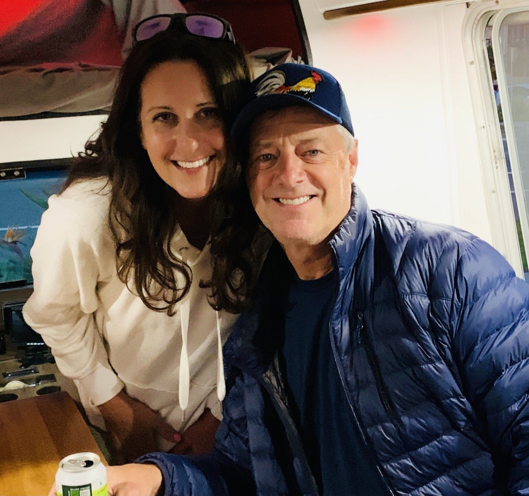 Smiling pair seated together in a small plane cabin, one in a white hoodie and one in a blue jacket and cap.