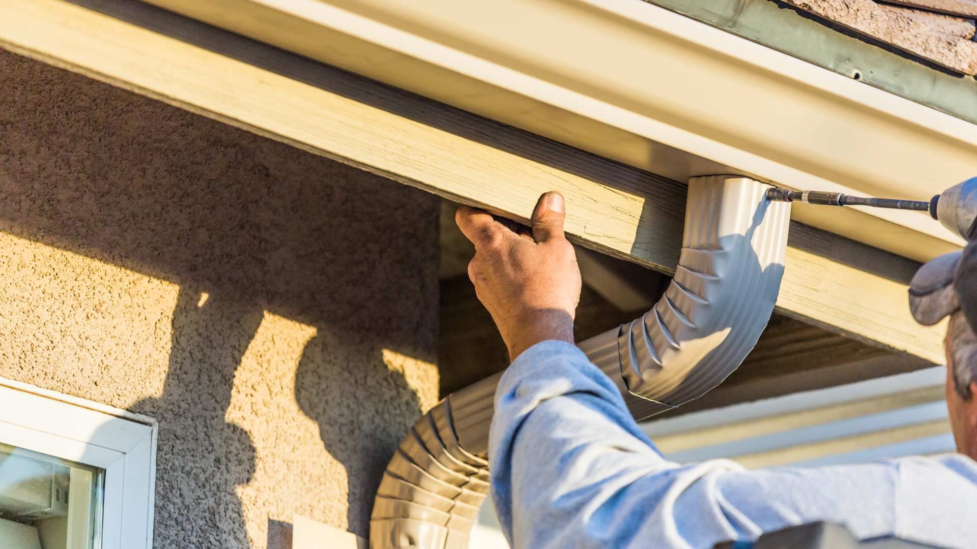 Worker installing a beige gutter on a house exterior with a drill and vent hose nearby
