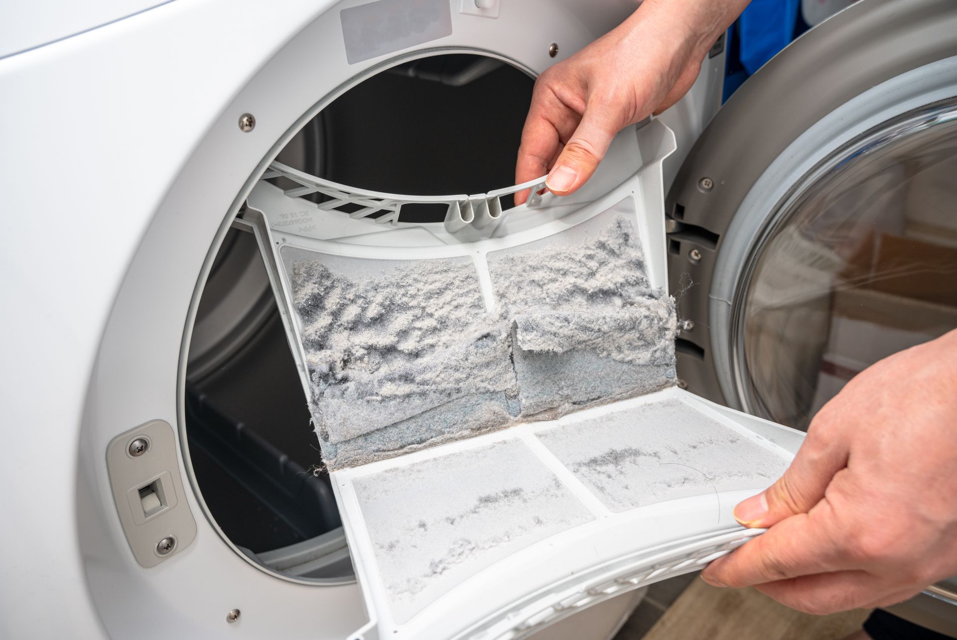 Person holding lint filter from dryer, full of grey lint, in front of open dryer door. Person holding lint filter from dryer, full of grey lint, in front of open dryer door.