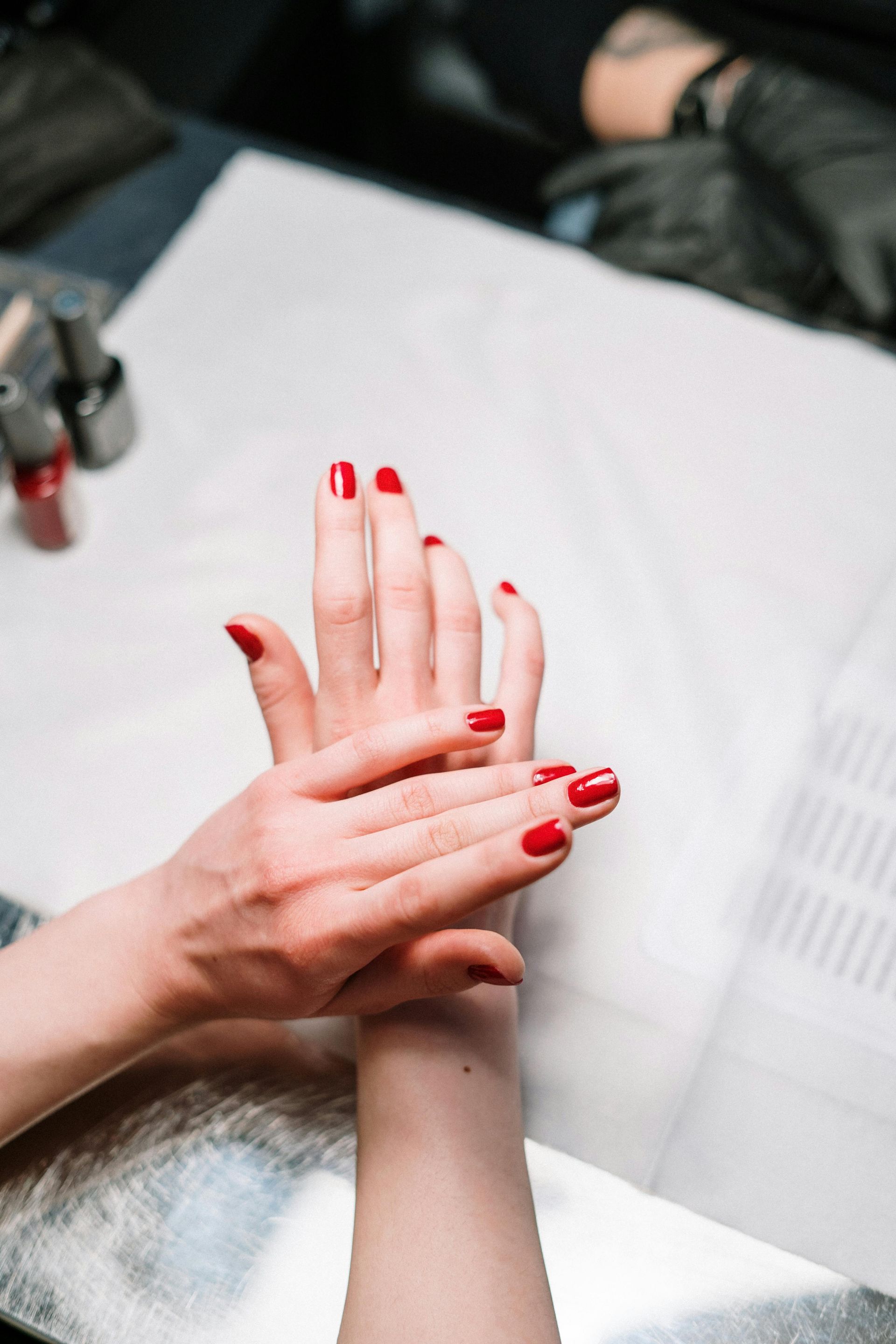 Mani con smalto rosso massaggiate su una superficie bianca, sullo sfondo una persona che indossa guanti neri.