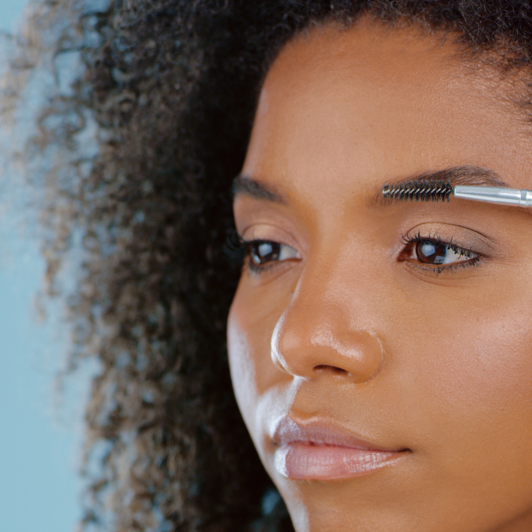 Woman grooming eyebrow with a brush, light blue background.