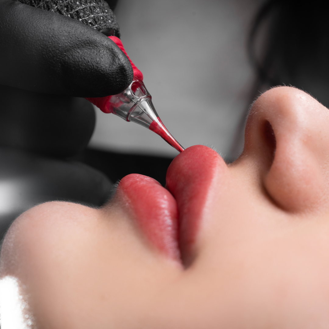 Person's lips being tattooed with red pigment by gloved hand holding a tattooing device.