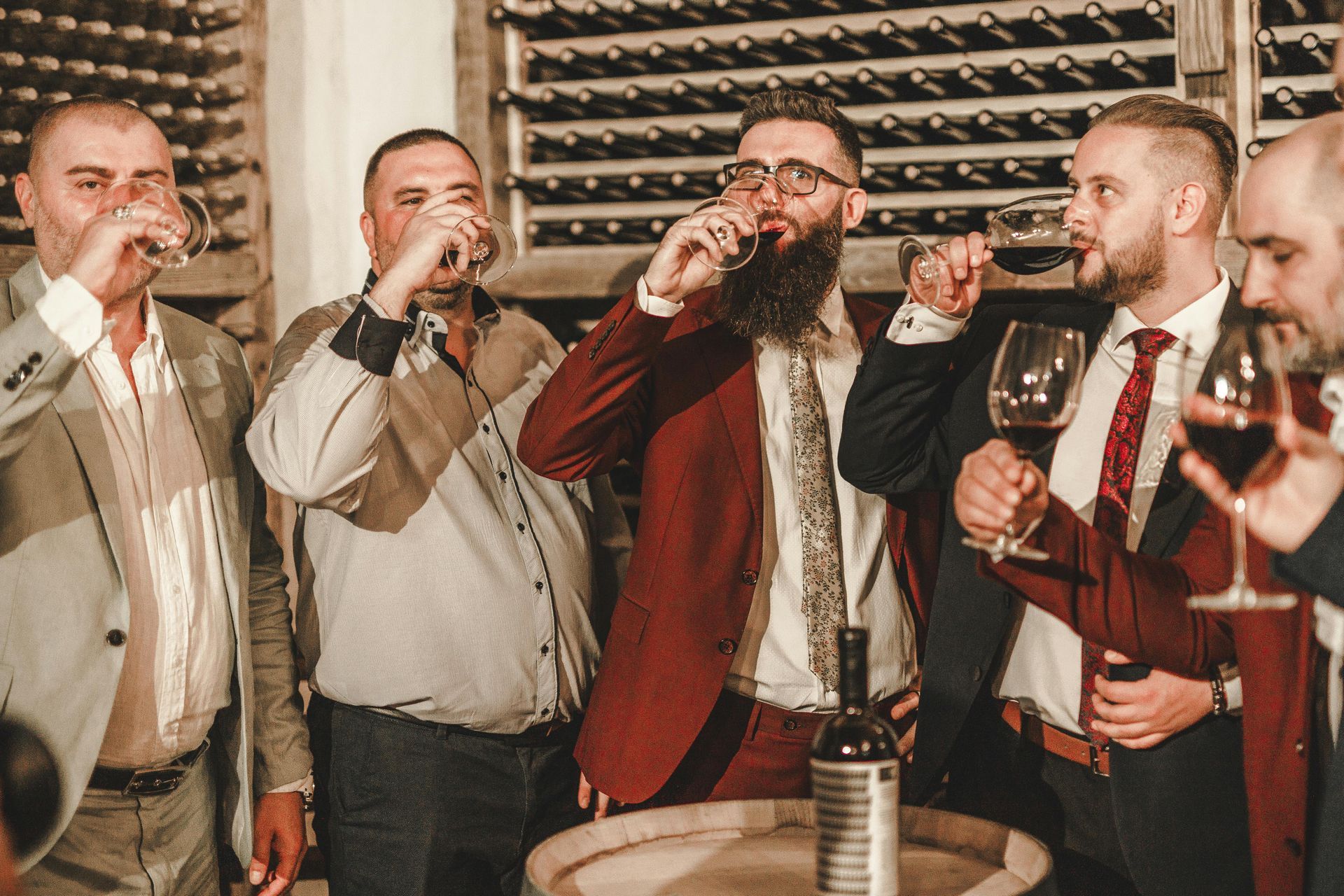 Men in suits tasting wine in a cellar; a wooden barrel and bottles are nearby.