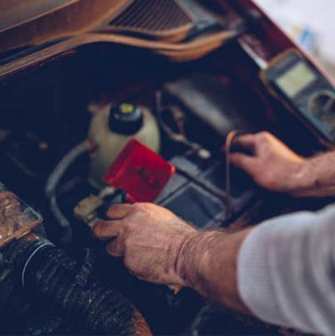 Hands working in a car engine bay, holding a red tool near components under the hood.
