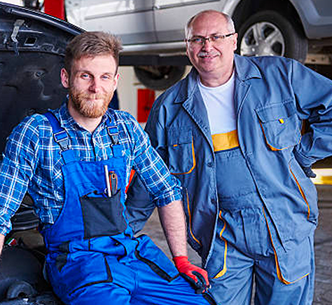 Two mechanics in blue coveralls standing beside a car in an auto repair shop, one wearing red gloves.