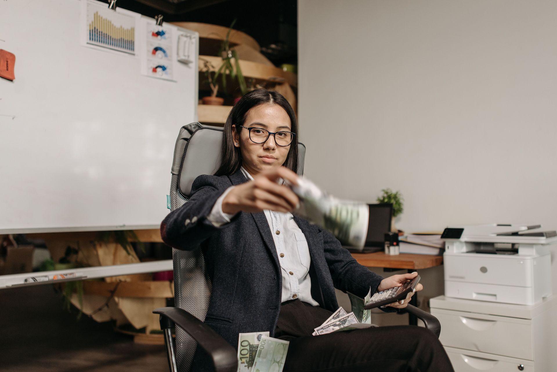 Person seated in an office, reaching toward the camera, with a whiteboard and printer in the background