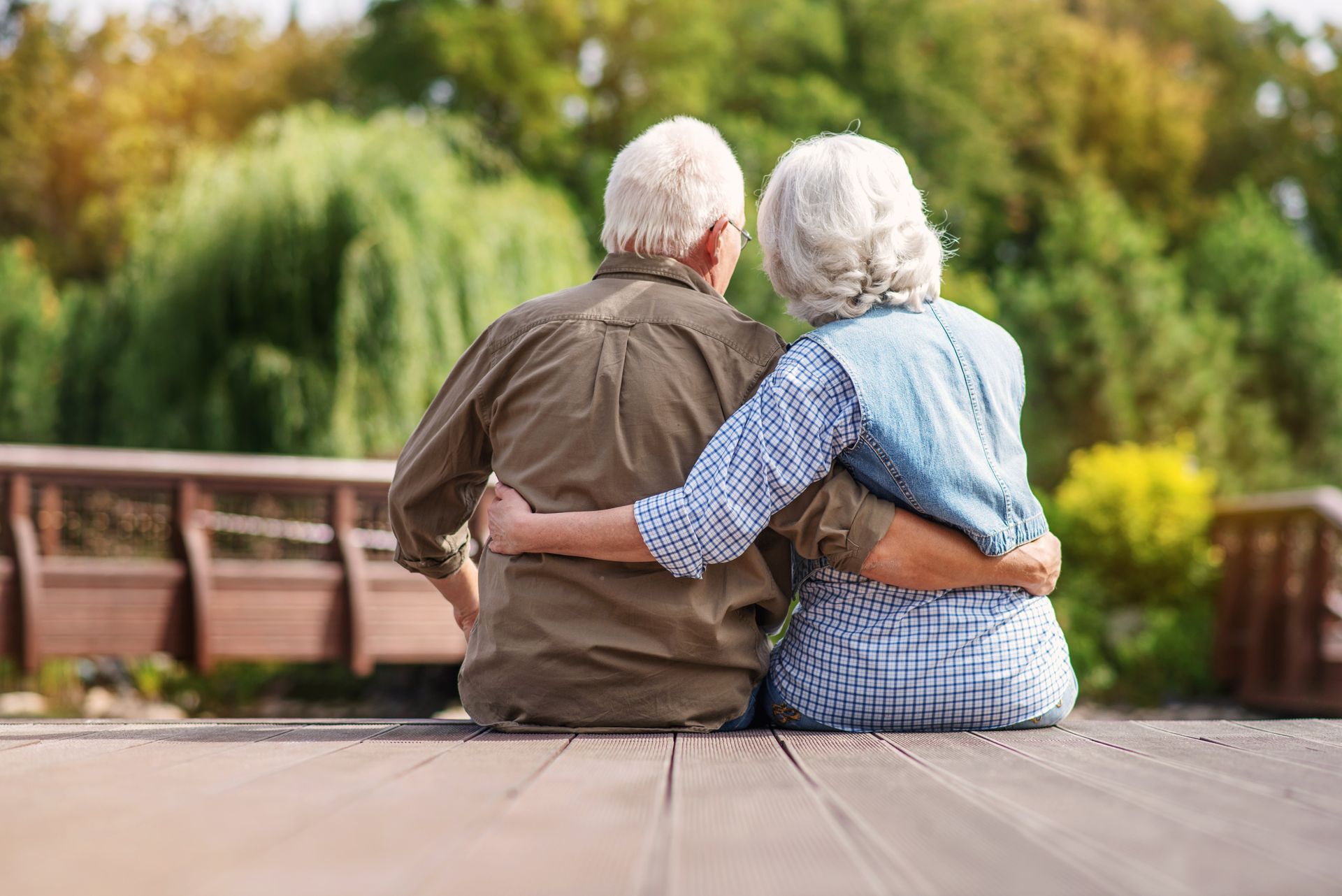 Elderly couple sitting on a wooden dock, embracing, looking out at a green landscape.