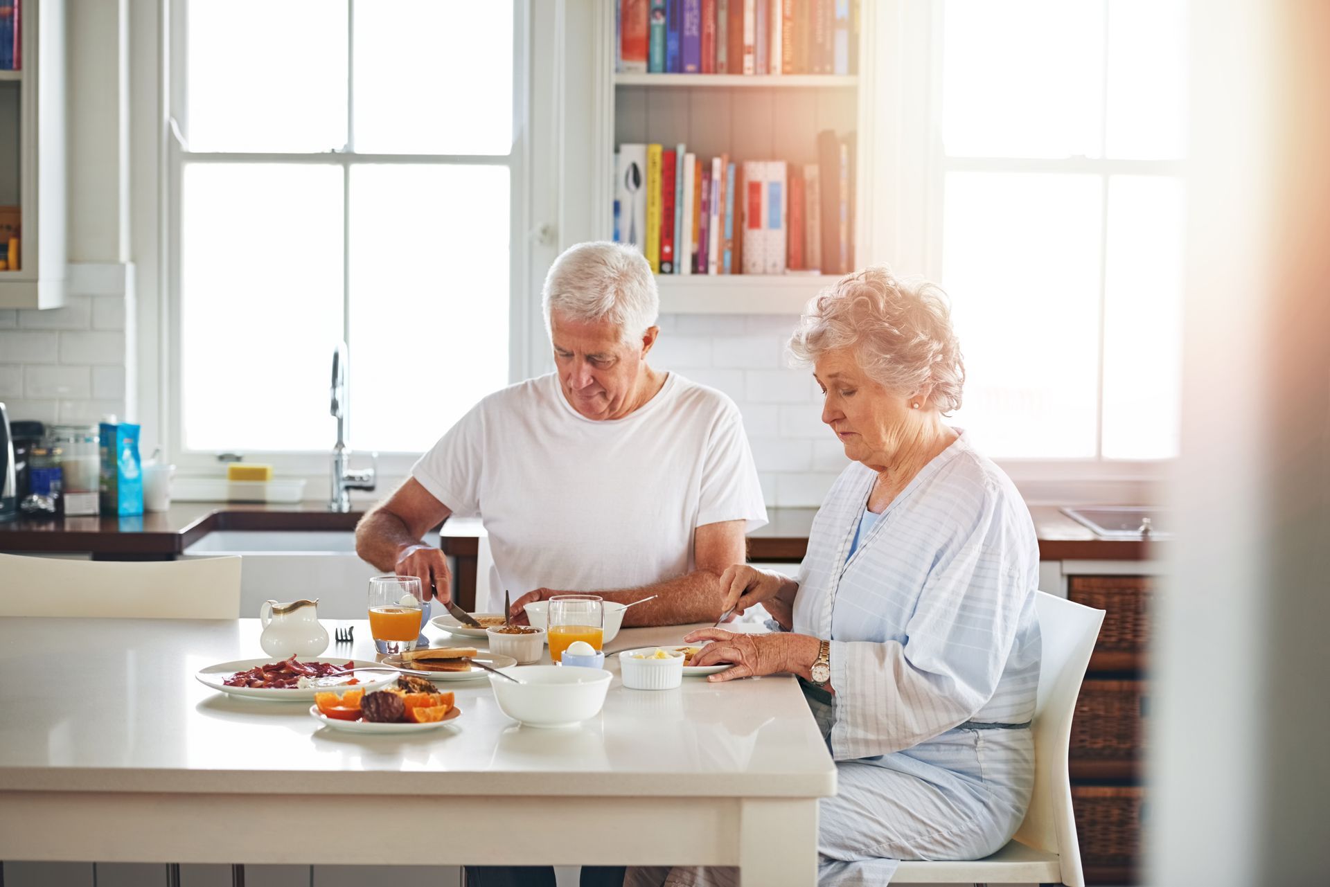 Elderly couple eating breakfast at a white kitchen table; natural light.