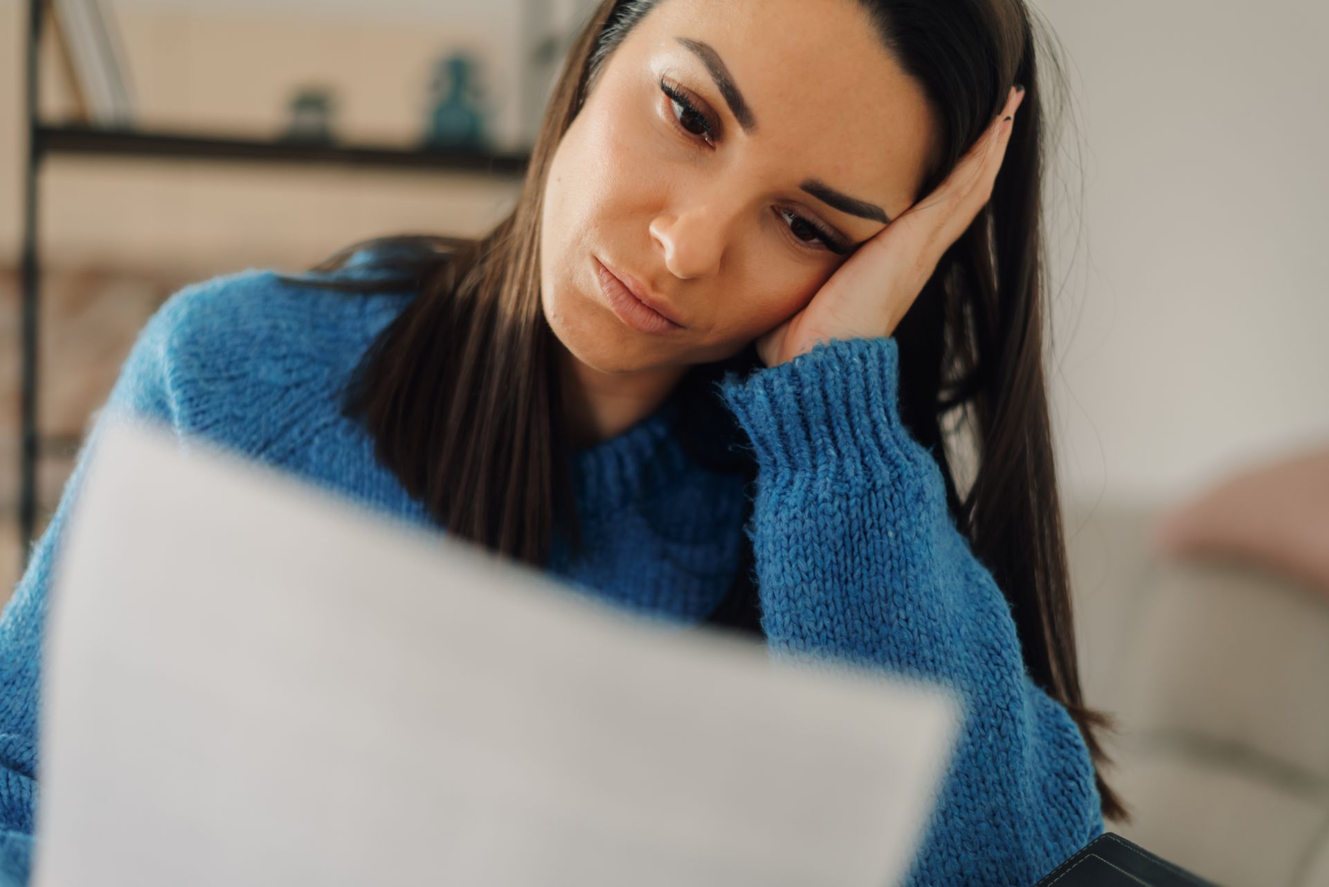 Woman looking at papers, appears worried, resting head on hand, wearing blue sweater.