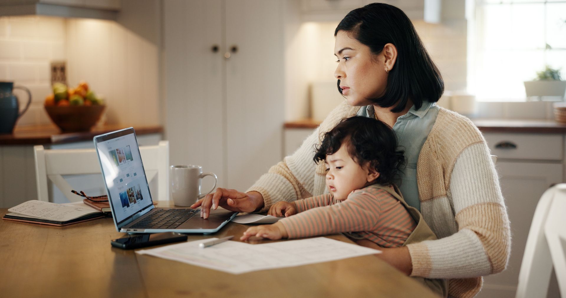 Woman works on laptop while holding a baby in a bright kitchen setting.