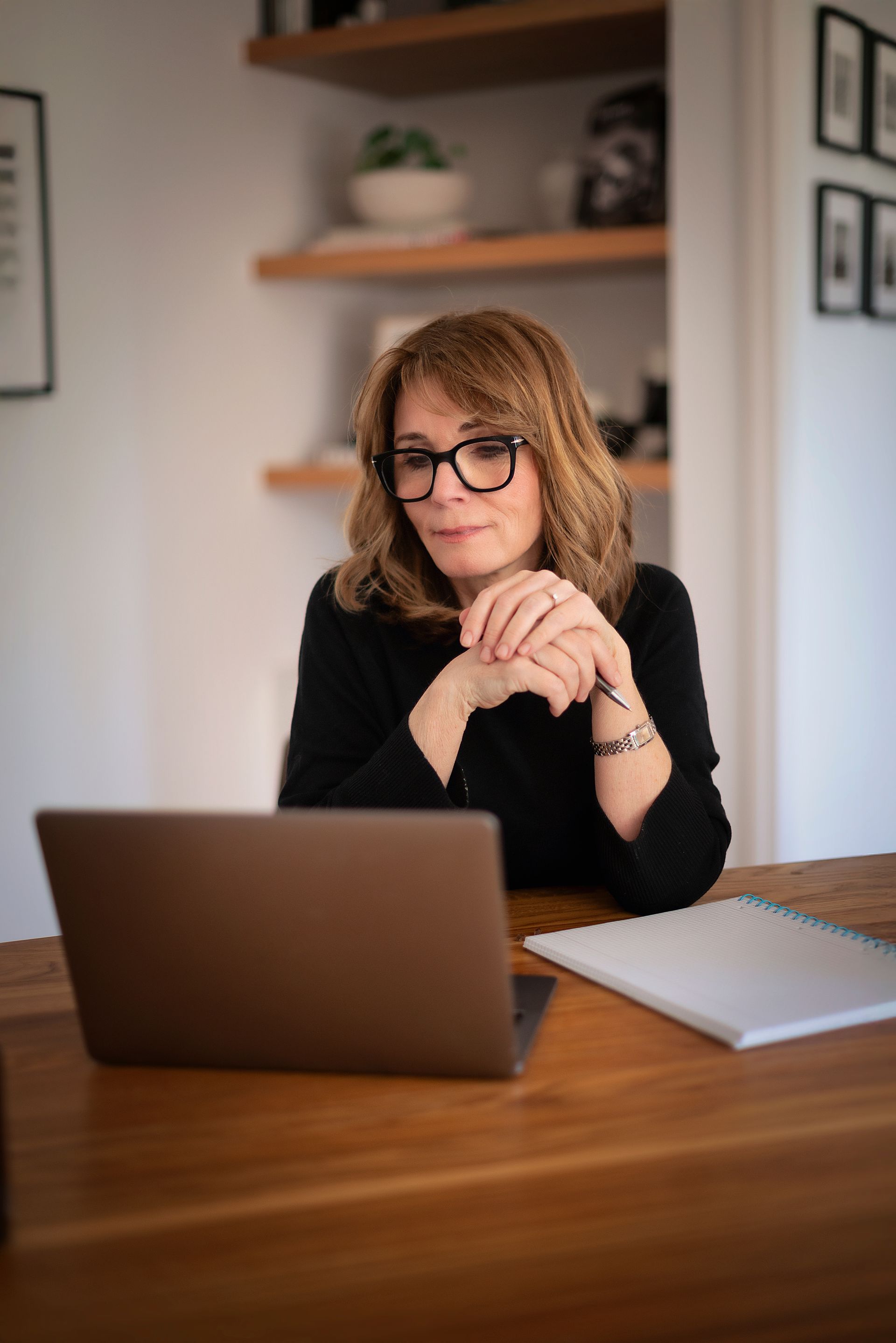 Woman with glasses sits at desk, working on laptop. Wooden desk with notebook.
