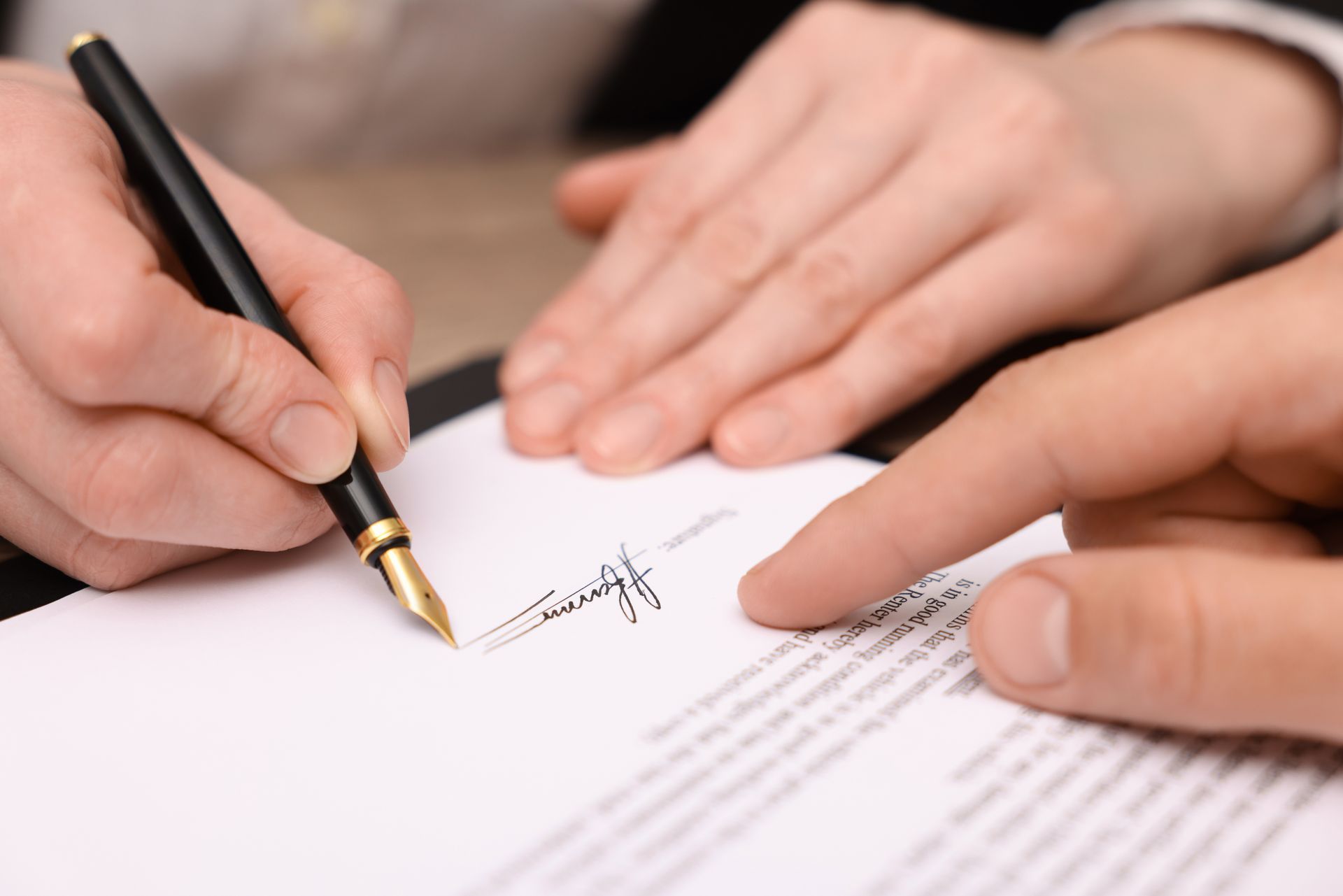 Person signing a document with a fountain pen, another person's finger pointing to the signature line.