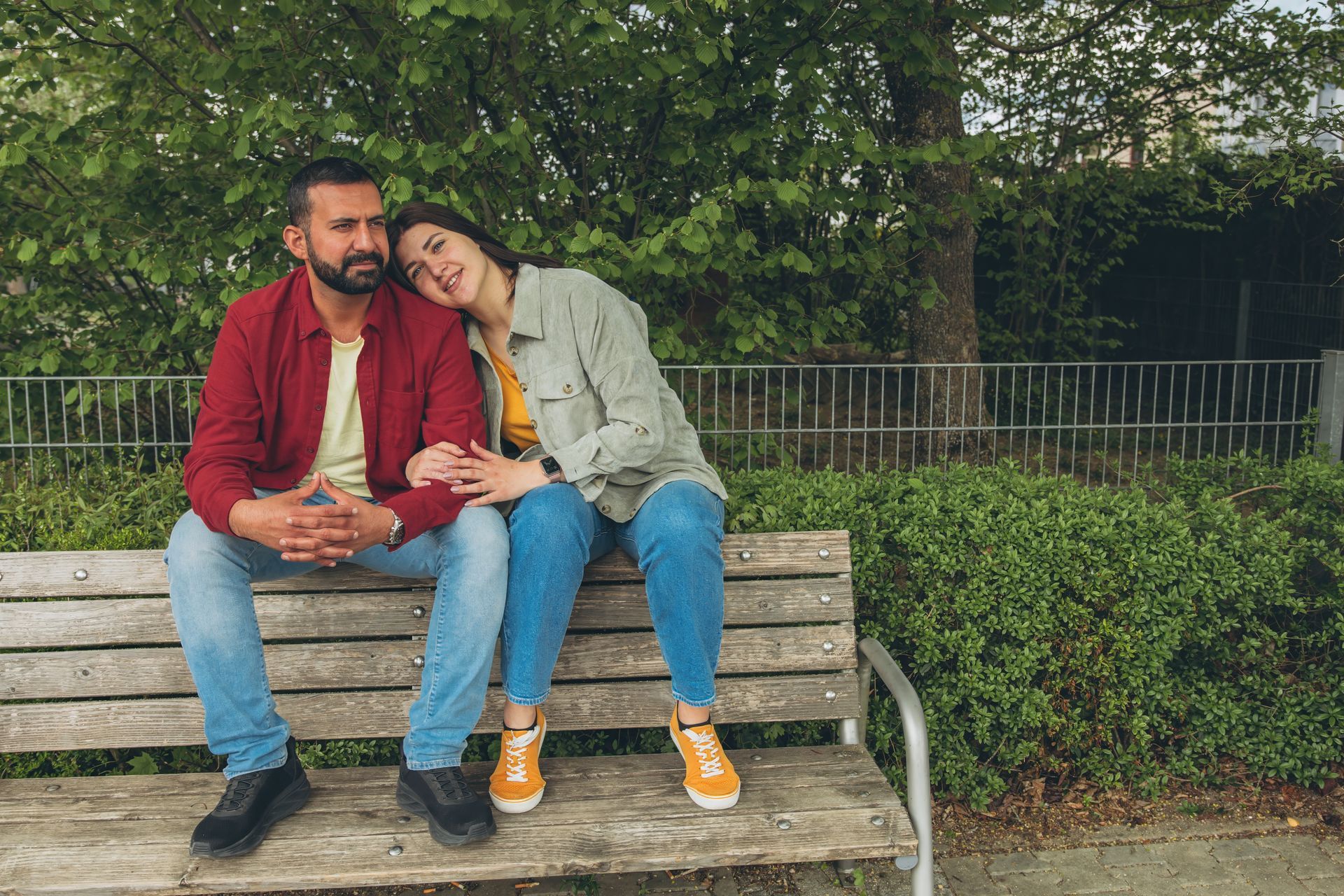 A couple sits on a bench. Woman rests head on man's shoulder. Park setting.