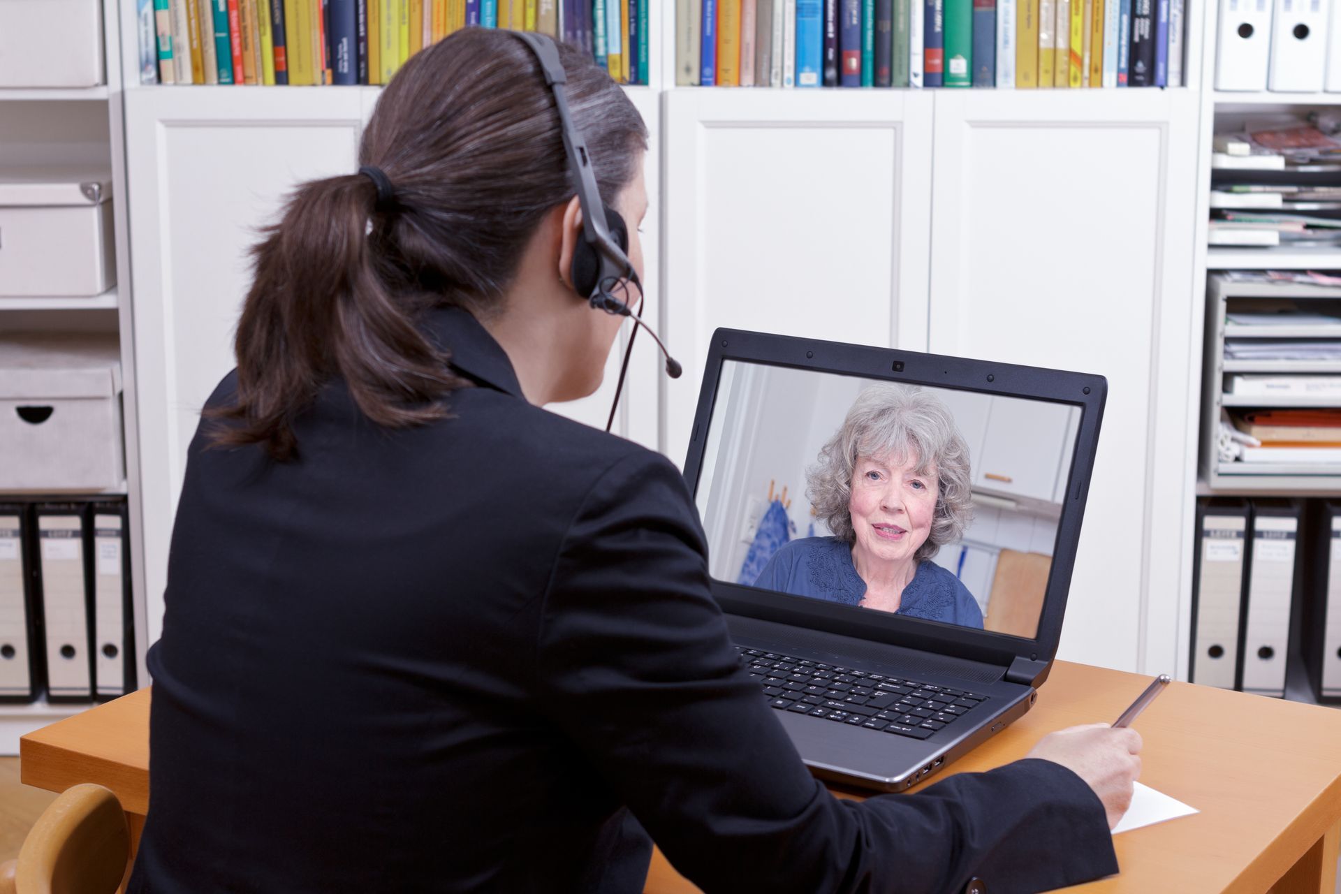 Woman in a blazer on a video call with a smiling older woman, taking notes, at a desk.