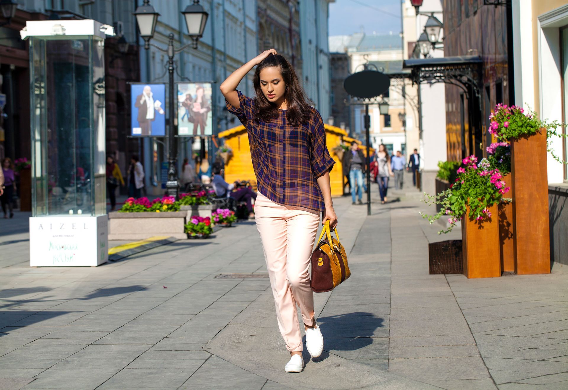 Woman walking down a city street, wearing a plaid shirt, pink pants, and carrying a bag.