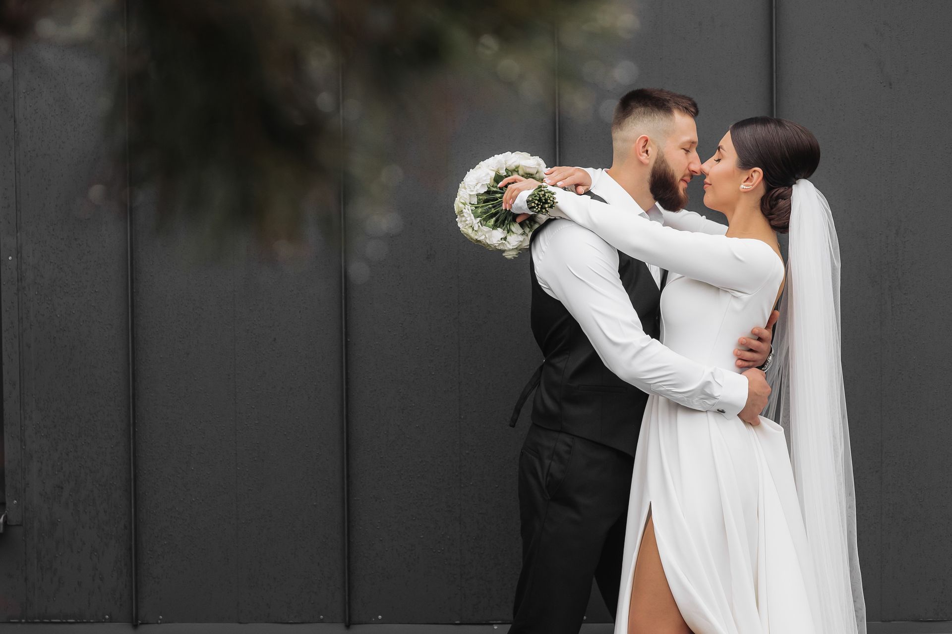 Newlyweds embrace outdoors; groom in black vest, bride in white dress, holding flowers.