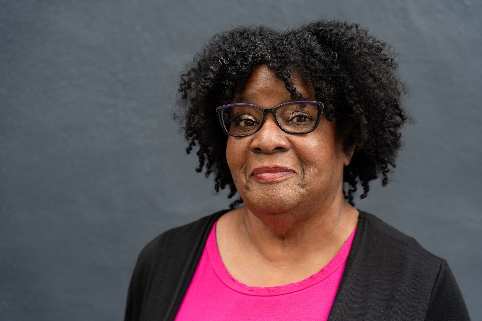 Woman with glasses and curly hair, wearing a pink shirt and black cardigan, smiling at the camera.