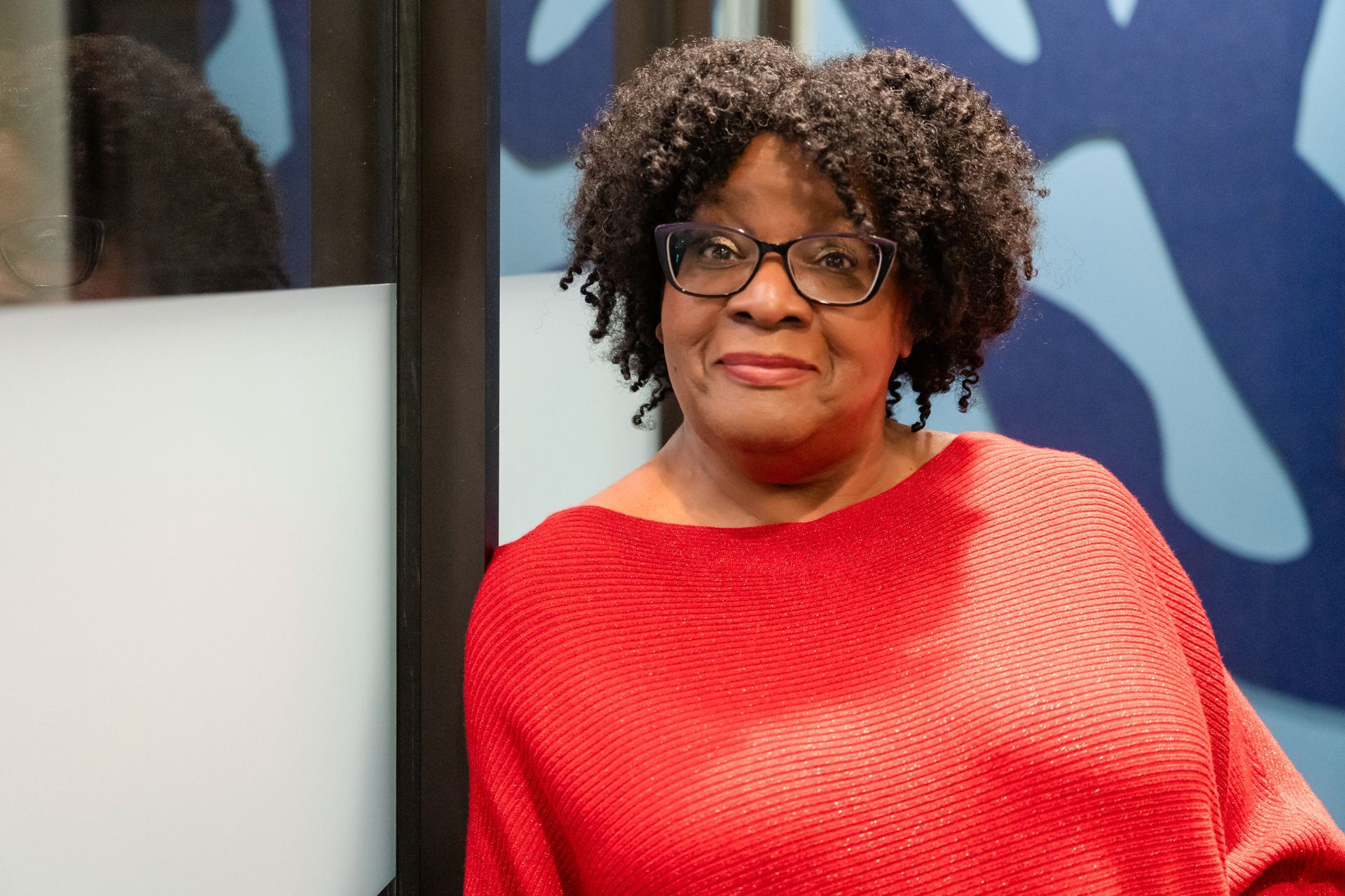 A woman wearing glasses and a red sweater is standing in front of a mirror.