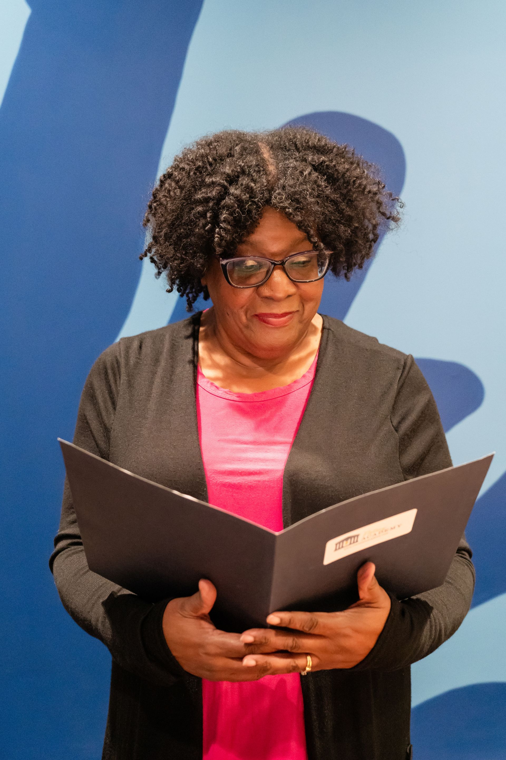 A woman wearing glasses is reading a book in front of a blue wall.