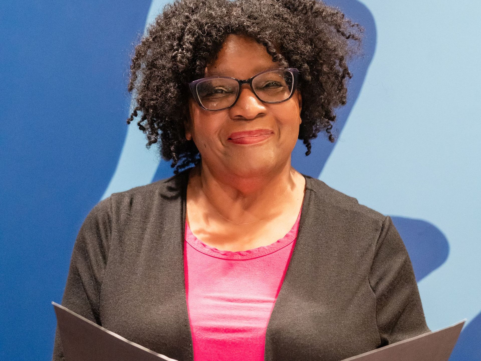 Woman with glasses smiling, holding papers. Wearing pink top, black cardigan. Blue background.