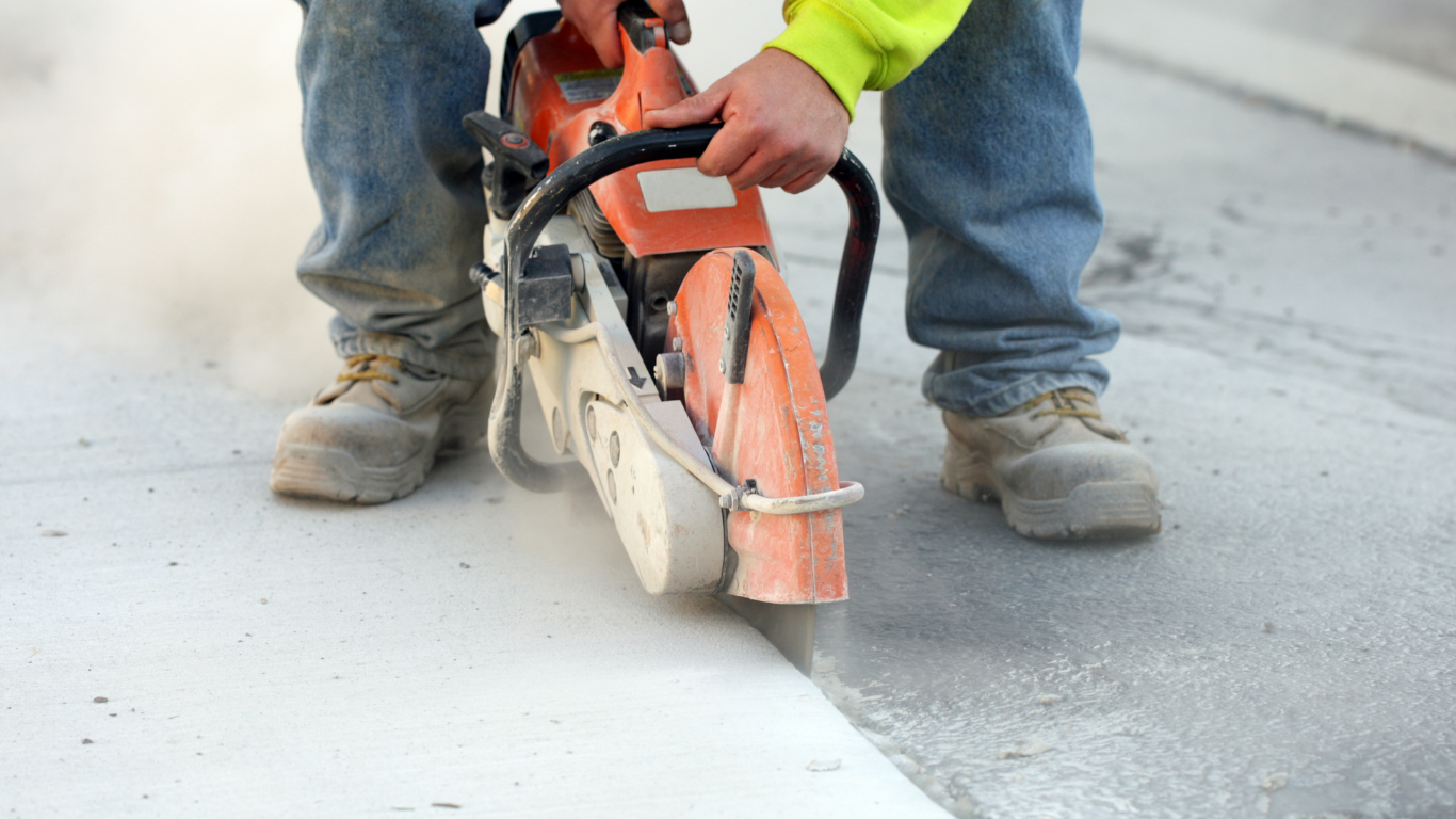 Person in work boots uses a concrete saw to cut a line in a sidewalk, creating dust.