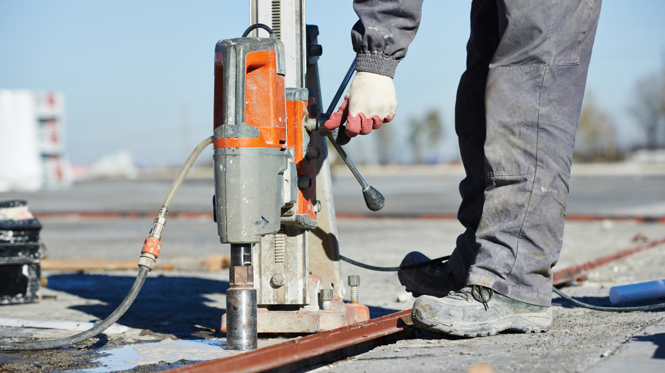 Worker using a drilling machine on concrete. Orange and gray power tool, blue jeans. Outdoors.
