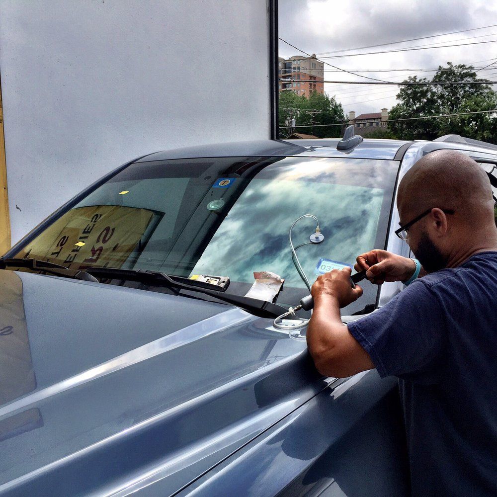 Man in navy shirt working on the hood of a gray car beside a building outdoors