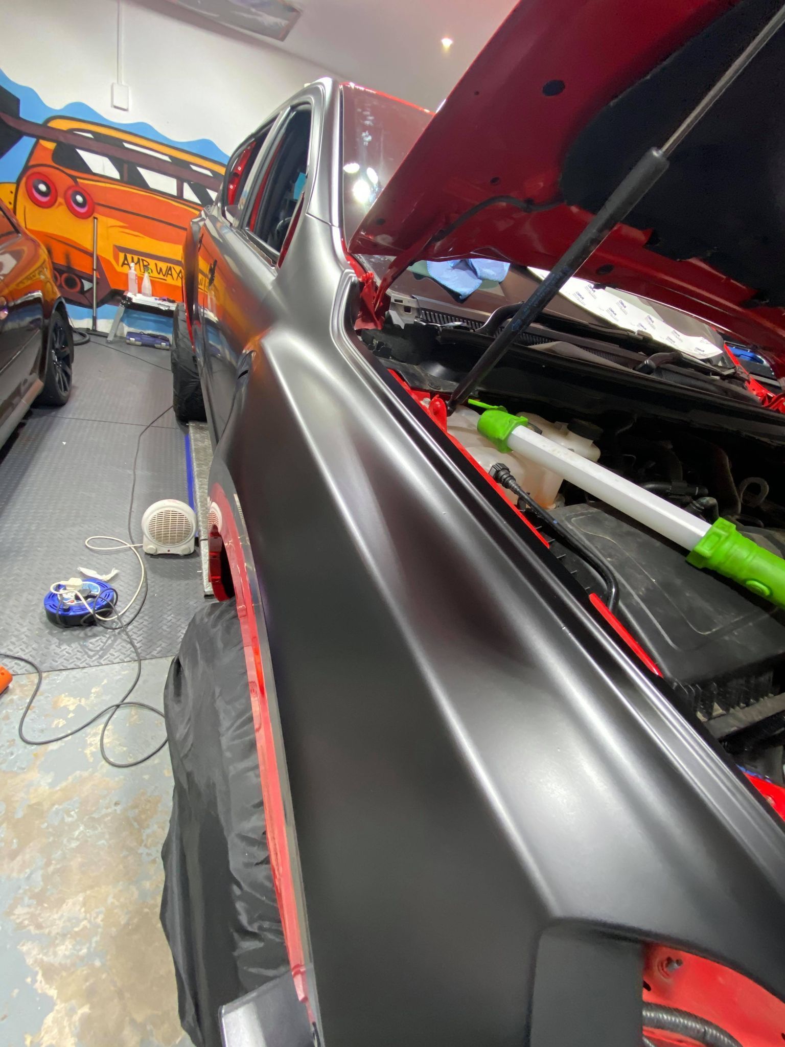 Black car in a garage with its hood open, beside an orange car and tools on the floor.