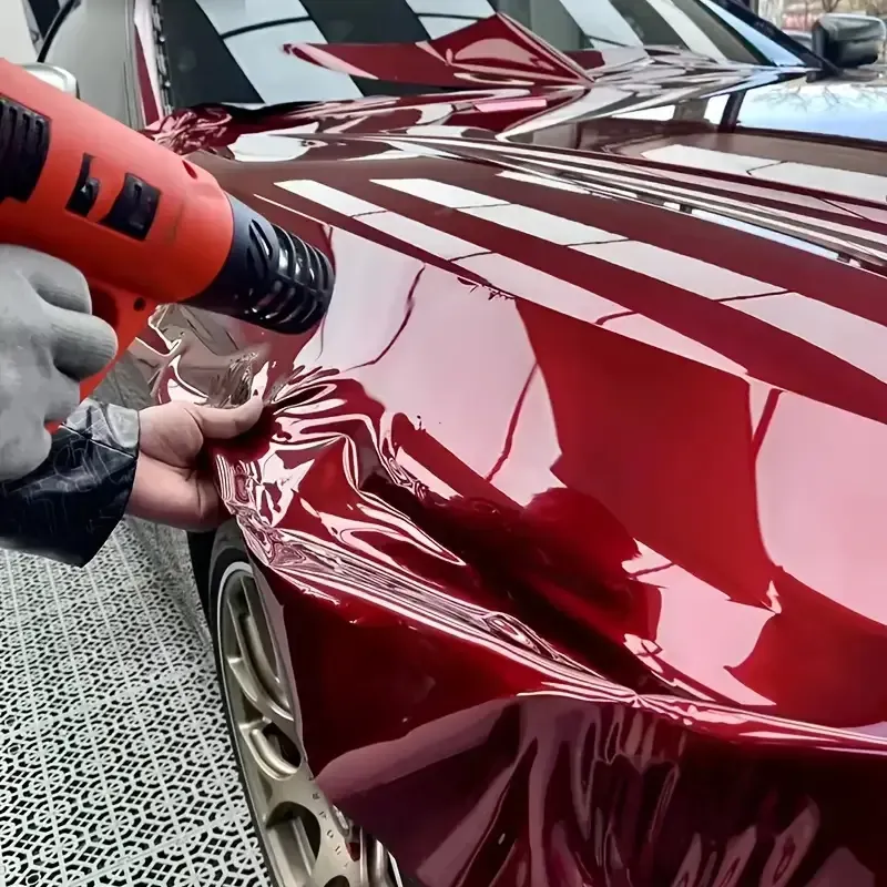 Person heat-guns wrinkled red car wrap on a shiny red car hood