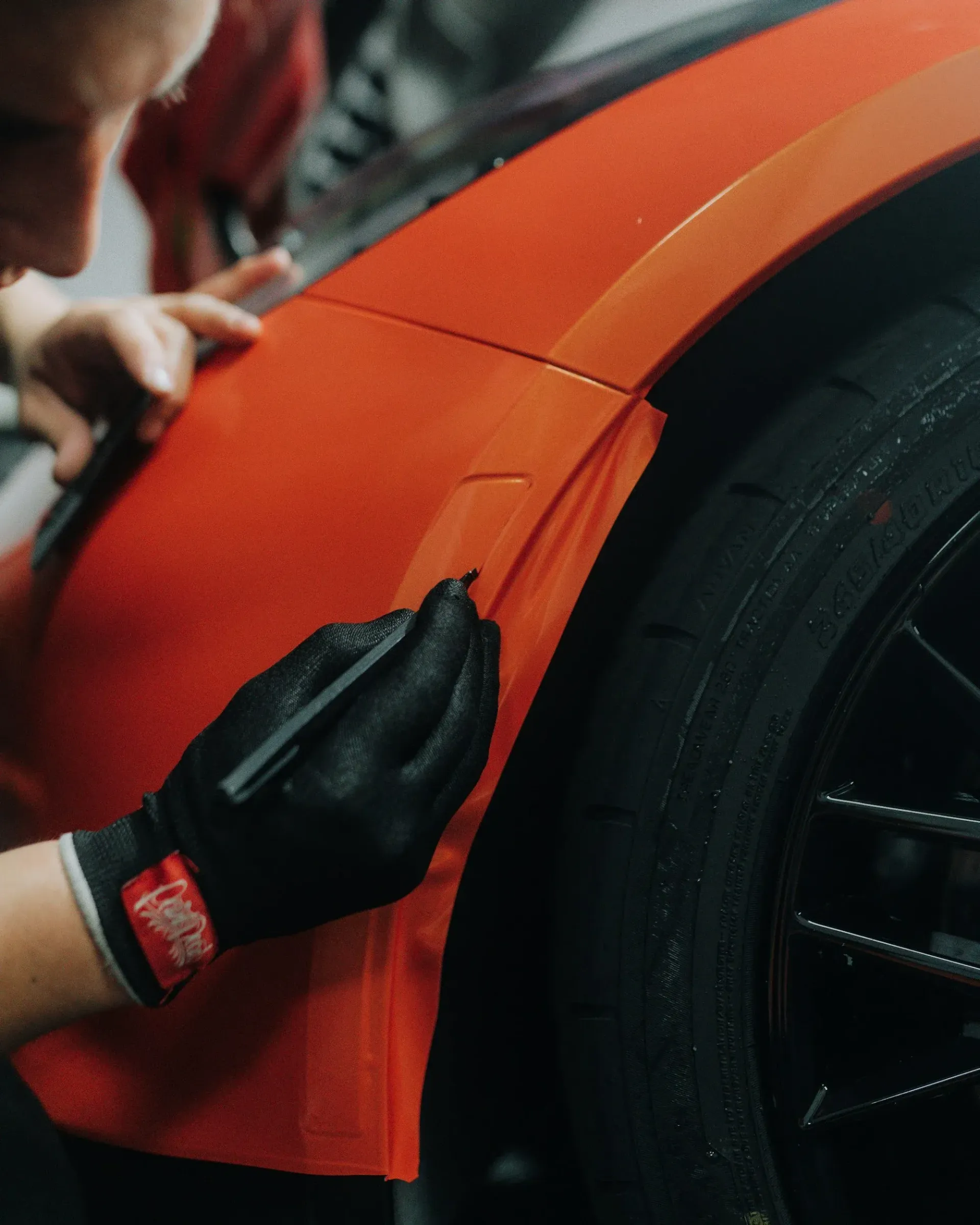 Gloved hands applying vinyl wrap to an orange sports car fender near the wheel