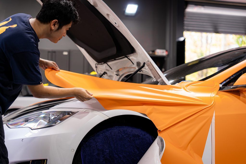 Person applying orange vinyl wrap to a white car in a garage