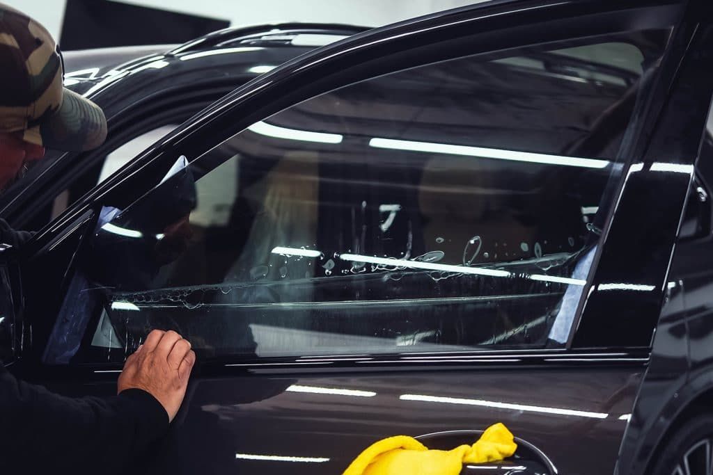 Person cleaning a black car window in a garage with bright overhead lights.