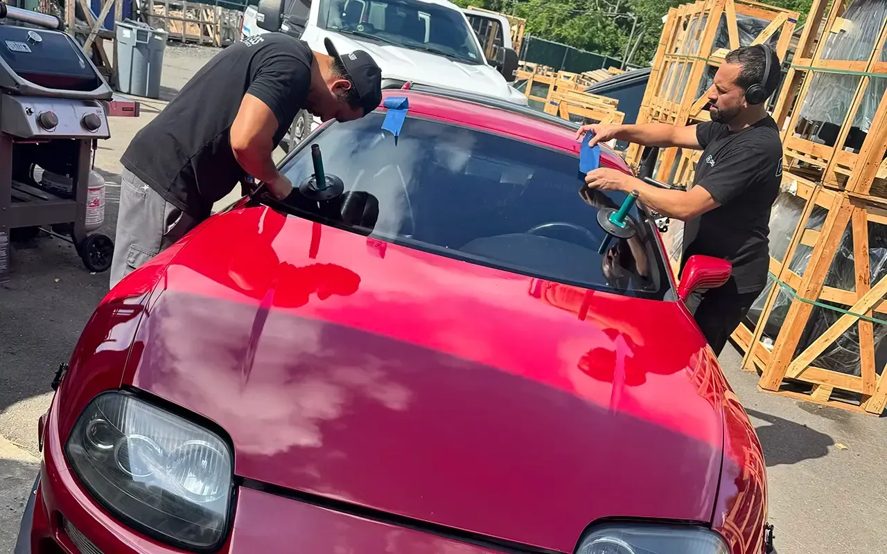 Two people wiping a red car’s windshield in a parking lot, with a tow truck in the background