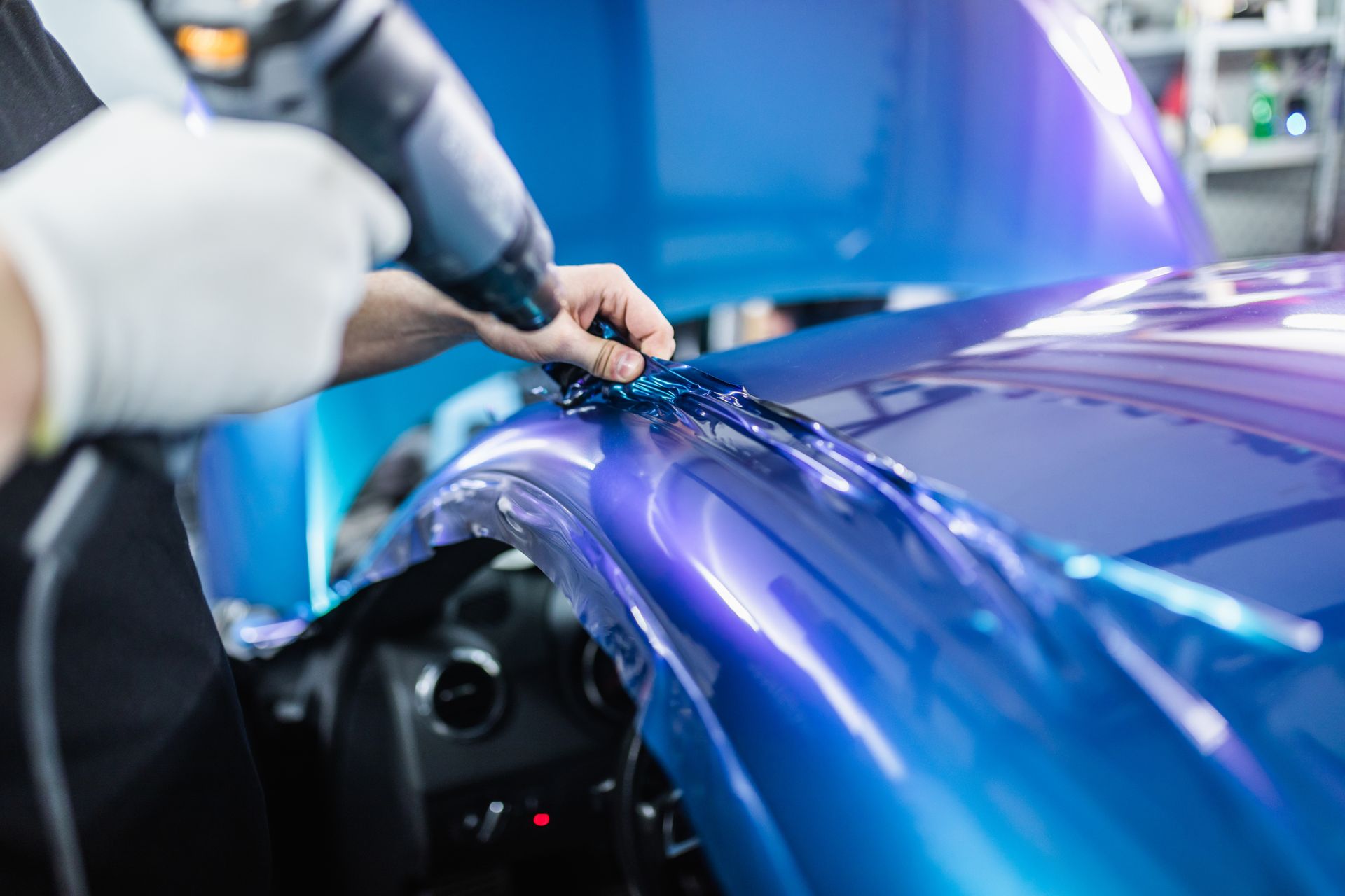 Hands applying protective film to a shiny blue car hood in an auto shop