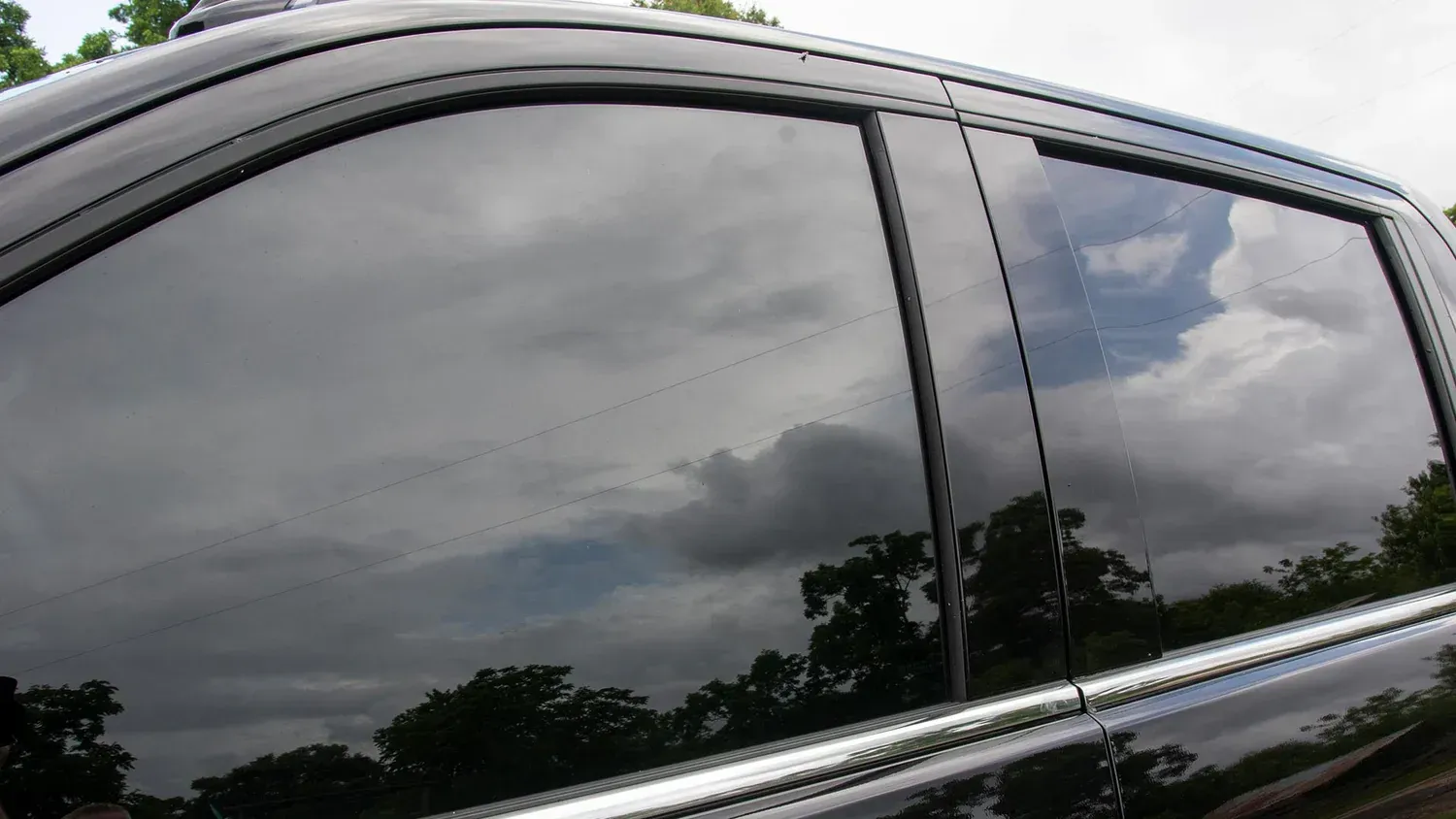 Dark-tinted car window reflecting cloudy sky and trees