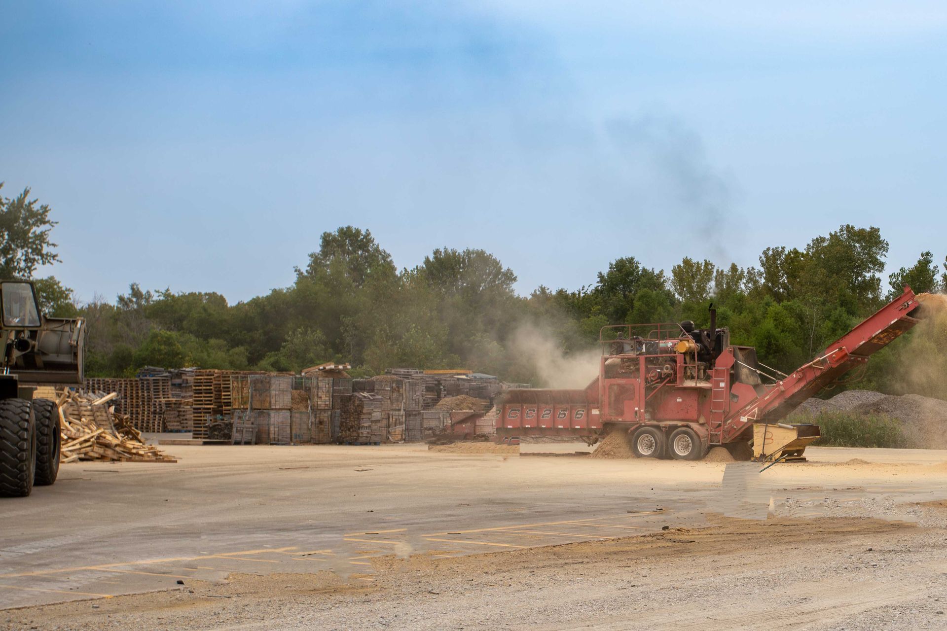 Industrial wood chipper blowing dust in a lumberyard, with stacked pallets and trees in the background.
