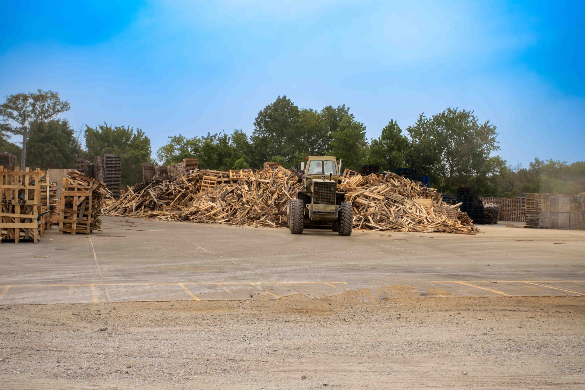 Tractor in a lumberyard, facing forward. Piles of wood and wooden crates are visible. Blue sky in the background.