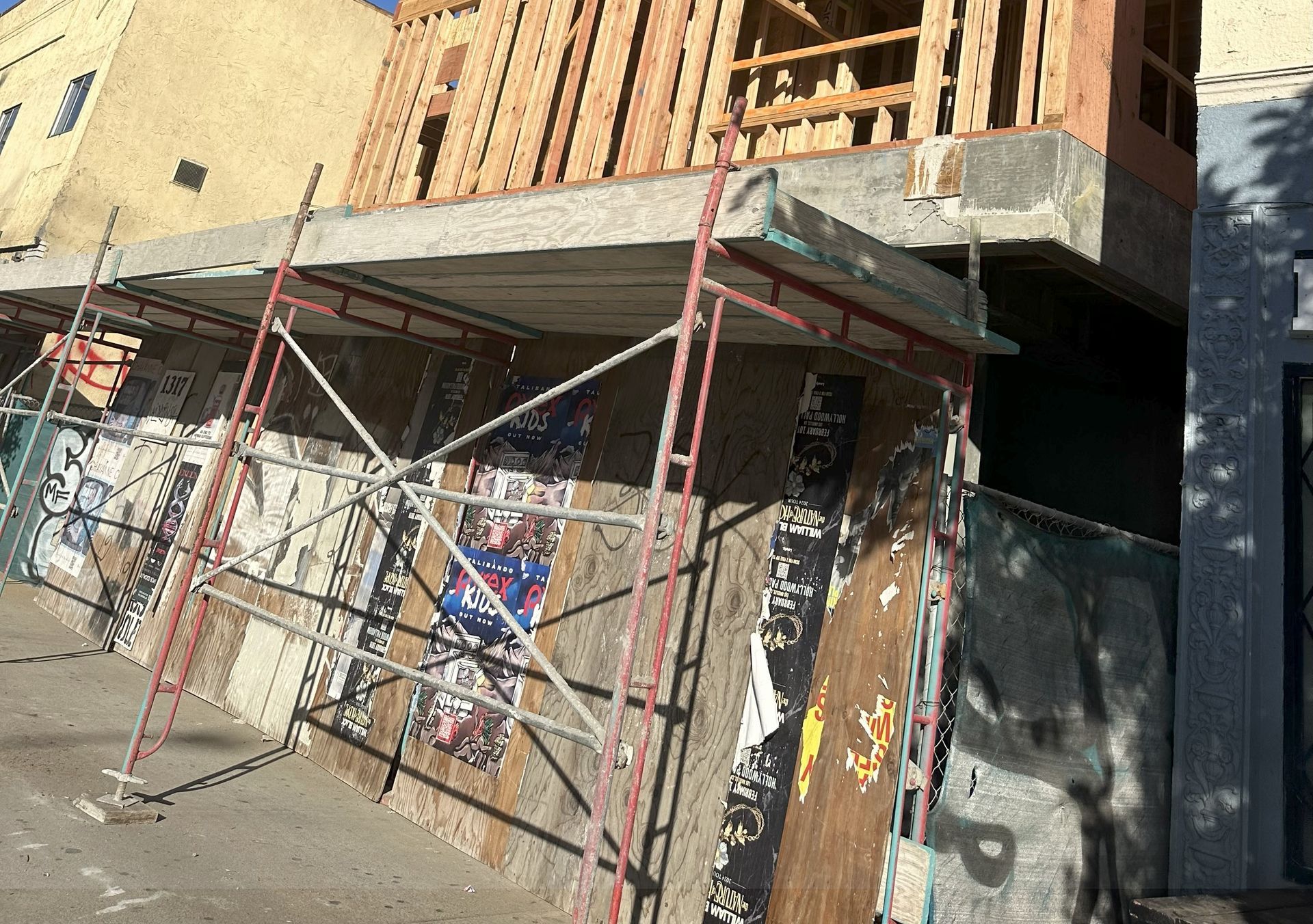 Scaffolding supports a concrete structure under construction on a building, against a wall covered in old posters.