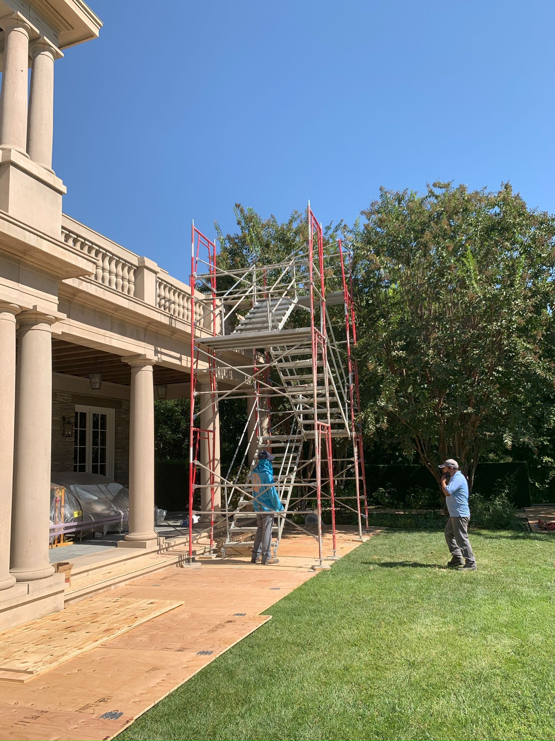 Scaffolding next to a building with columns; two people near it, one on lawn. Sunny day.