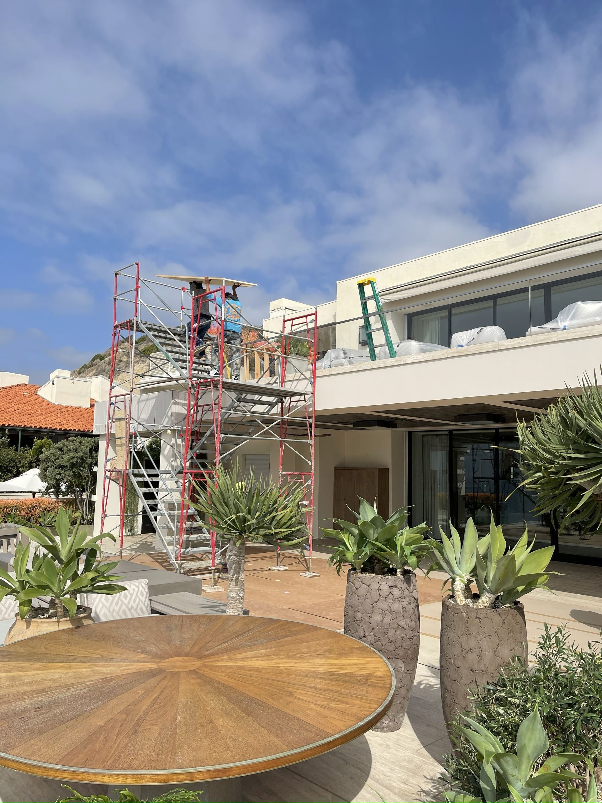 Construction on a building, with scaffolding, a ladder, and plants in front. Blue sky.