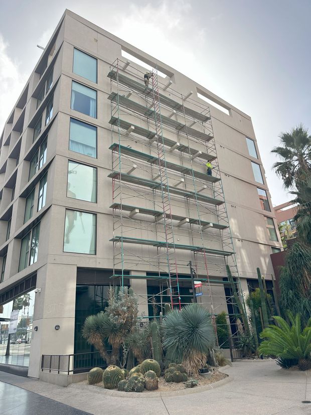 Scaffolding on the side of a beige multi-story building; a person works on scaffolding near windows, some plants in front.