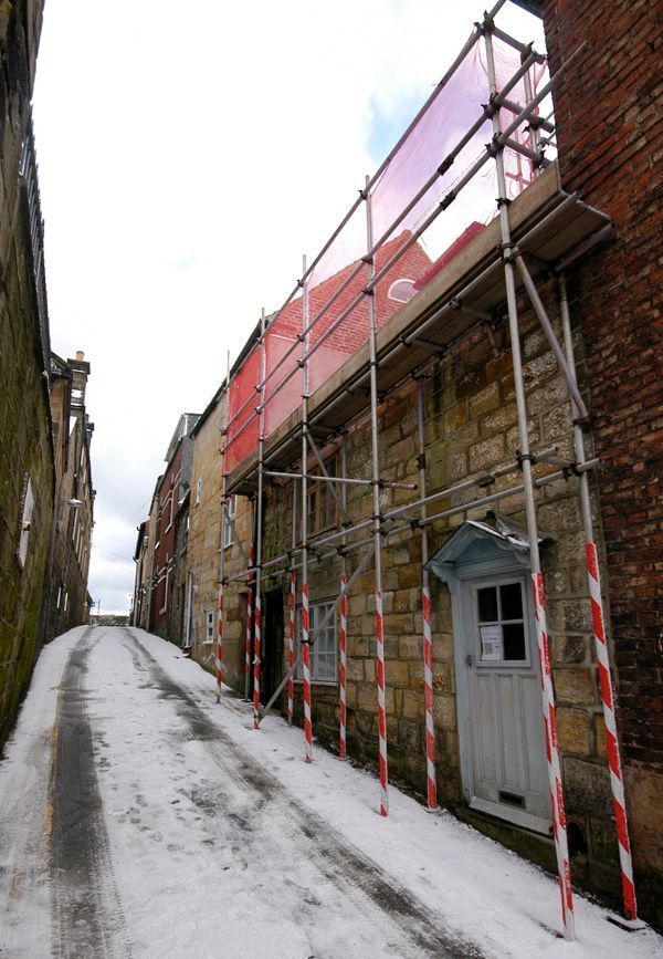 Snowy alley with a stone building under scaffolding; red safety netting; white door.