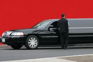 Black limousine with a suited chauffeur standing beside it on a red background.