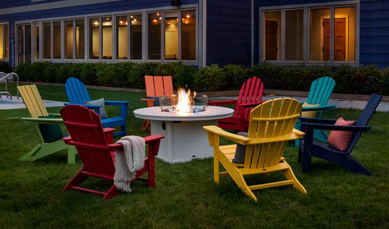 Colorful Adirondack chairs circle a lit fire pit on a grassy lawn.