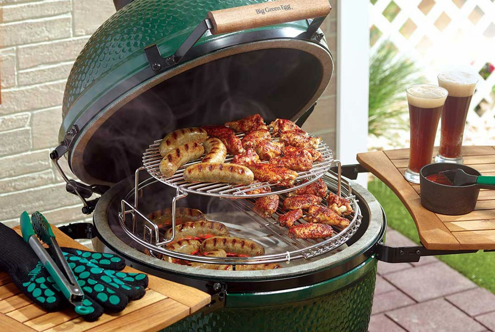 Green Big Green Egg grill with food and beer on a patio.