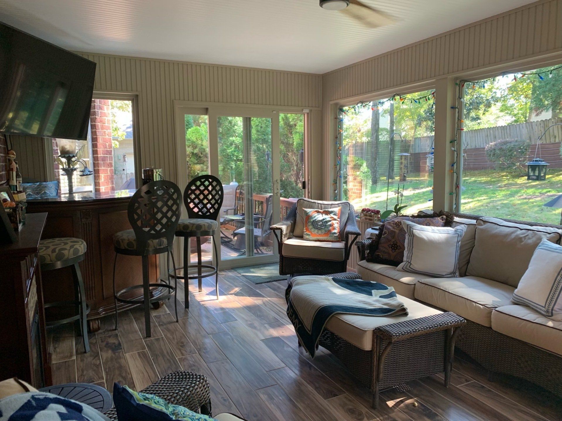 Sunroom with wicker furniture, bar stools, and large windows overlooking a yard.