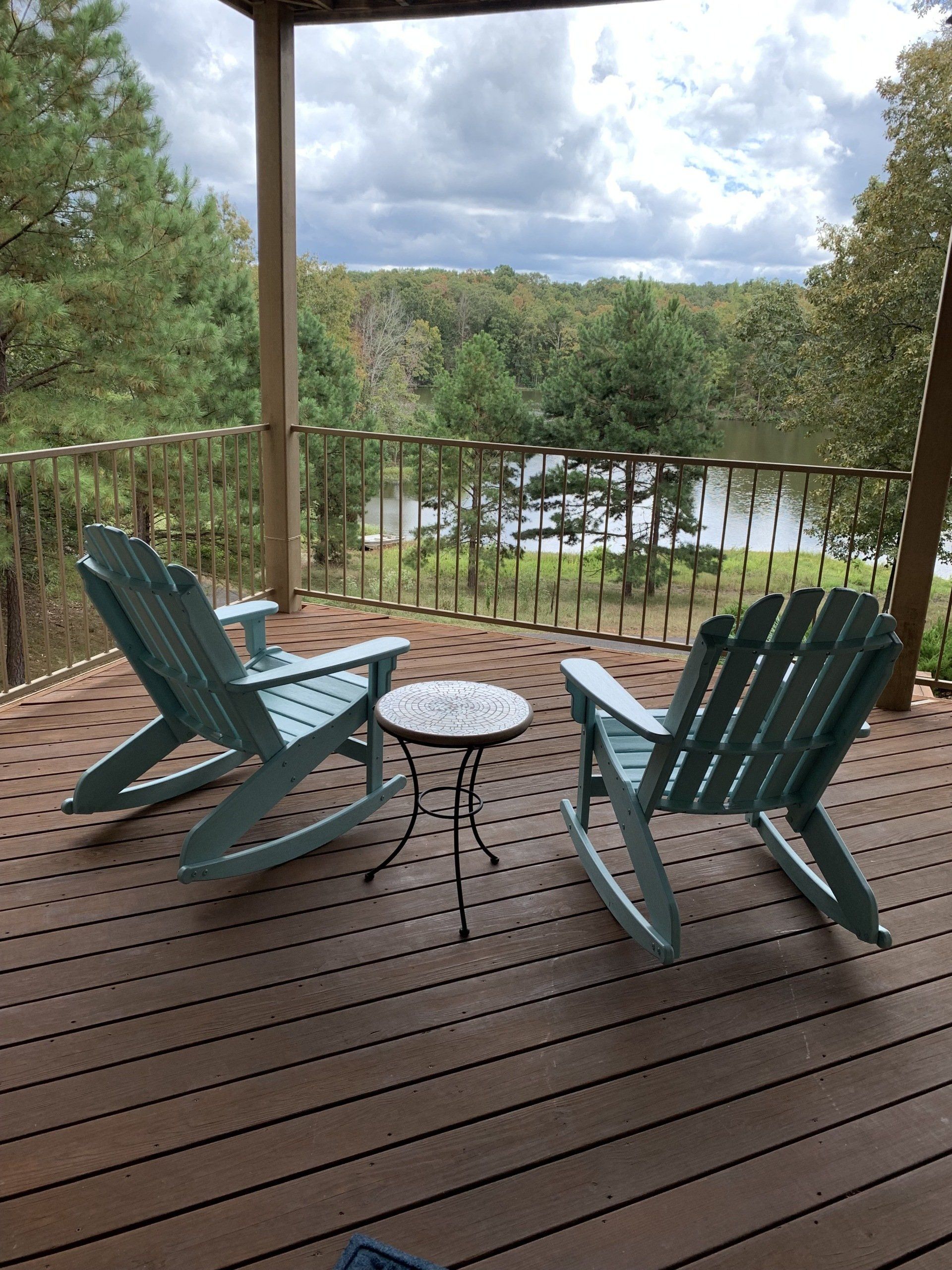 Two blue rocking chairs and small table on a wooden deck overlooking a lake and trees.