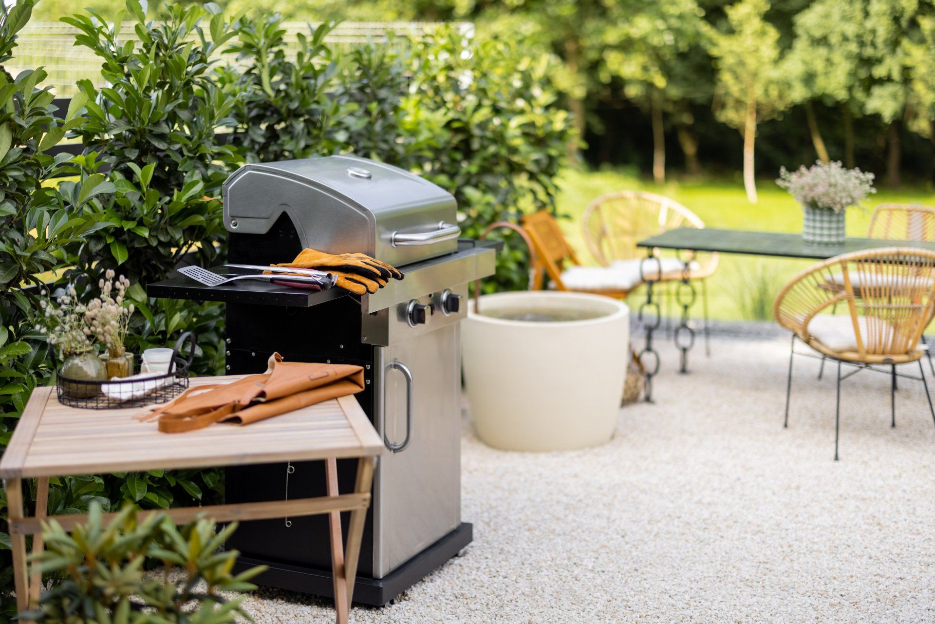 Grill on patio with table, wicker chairs, and greenery; sunny outdoor scene.